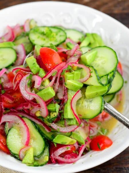 Cucumber tomato onion salad with avocado and fresh herbs being scooped from a bowl with a serving spoon.