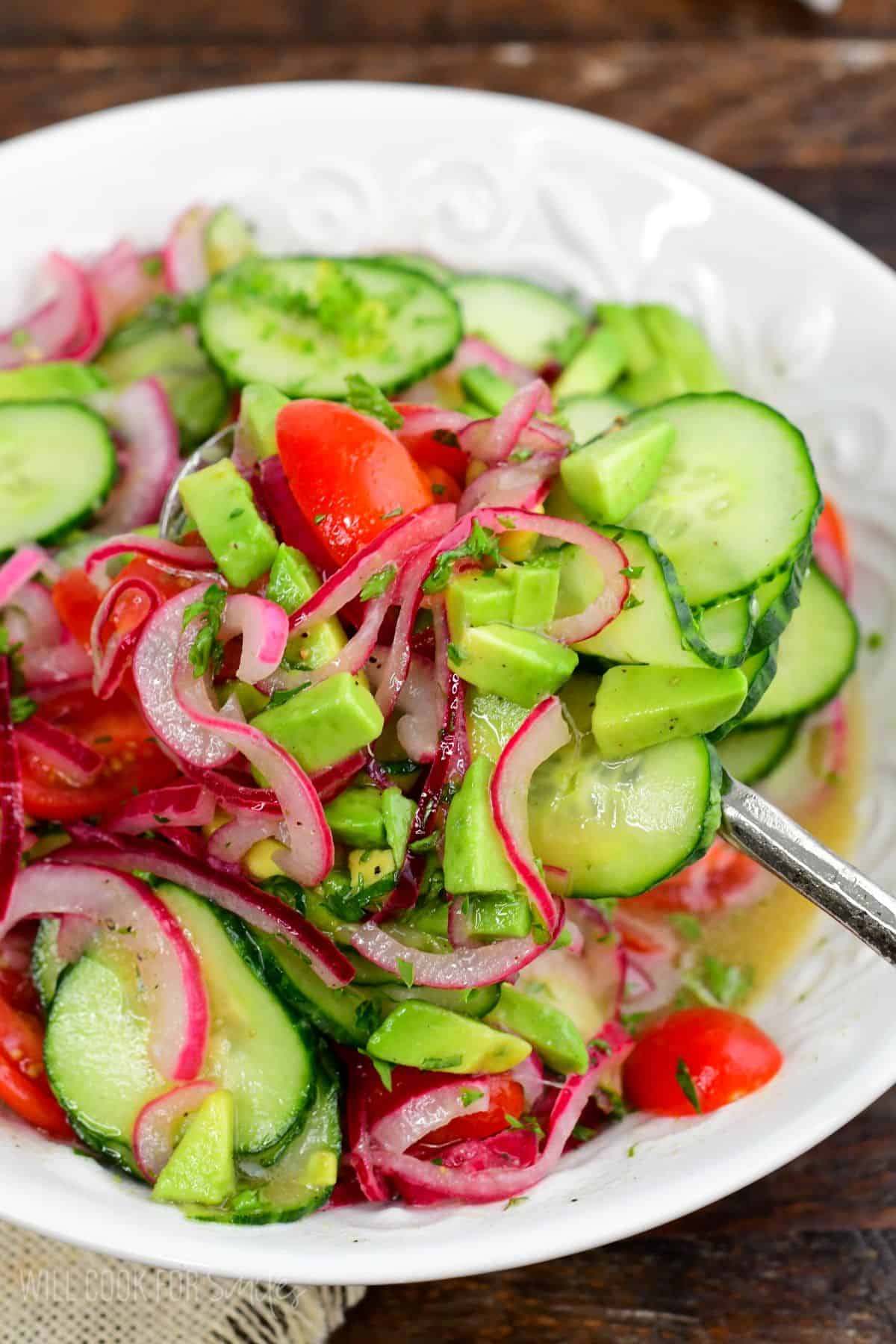 Cucumber tomato onion salad with avocado and fresh herbs being scooped from a bowl with a serving spoon.