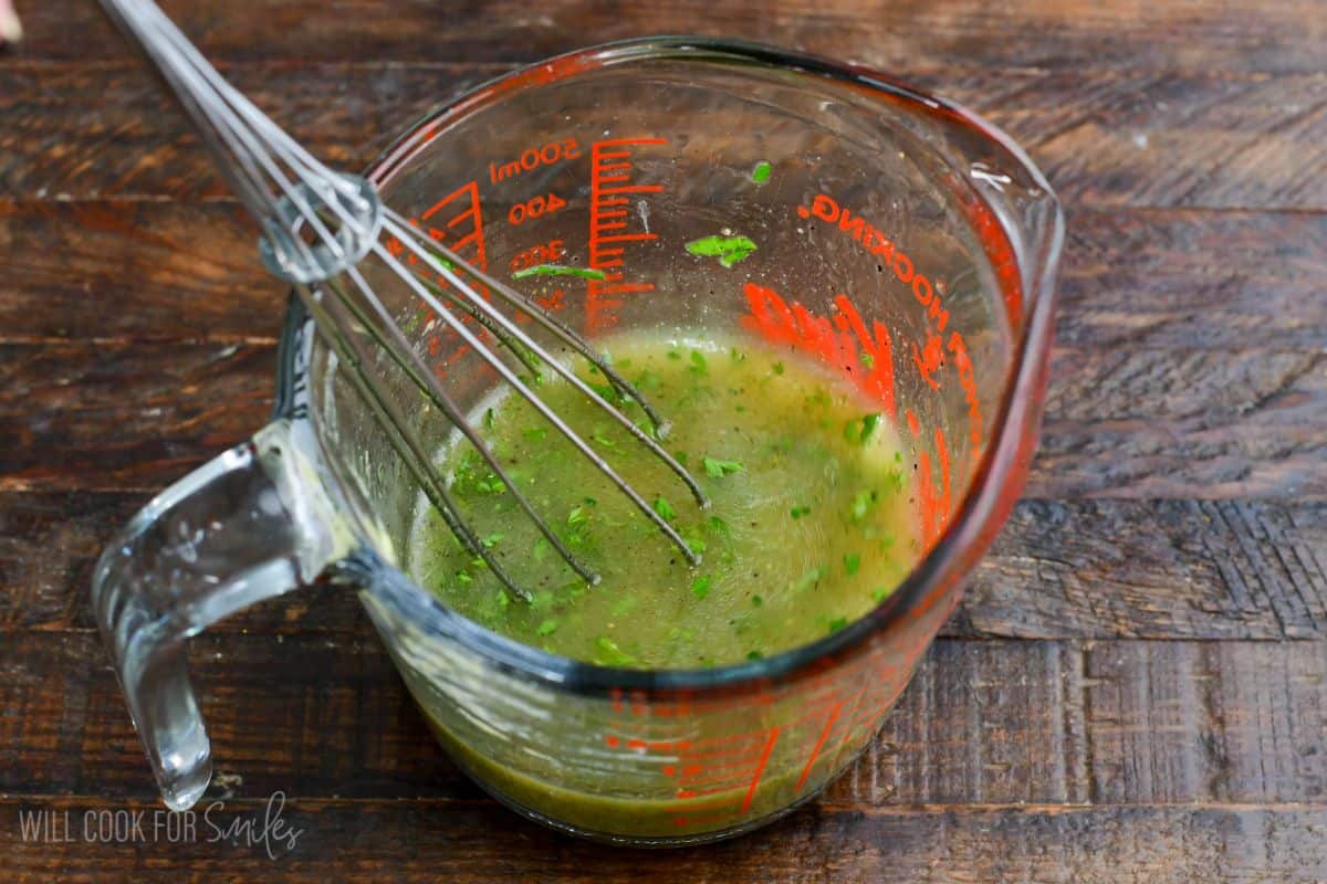 Homemade cucumber tomato salad dressing with fresh herbs being whisked in a glass measuring cup.