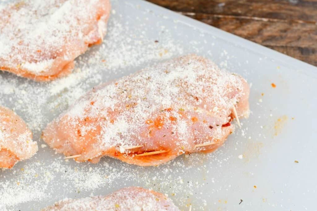 Stuffed chicken breasts coated with grated Parmesan before cooking on a cutting board.