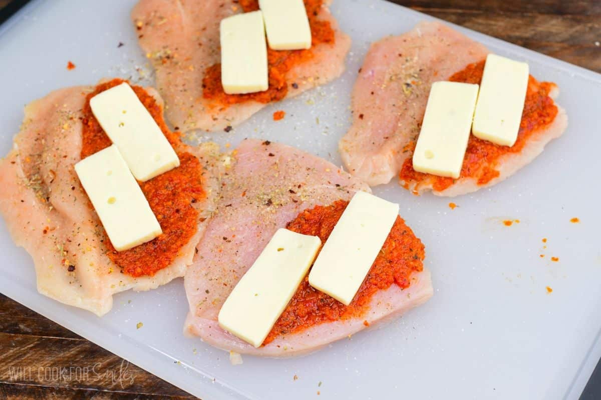 Butterflied chicken breasts topped with sun dried tomato pesto and slices of fontina cheese on a cutting board.