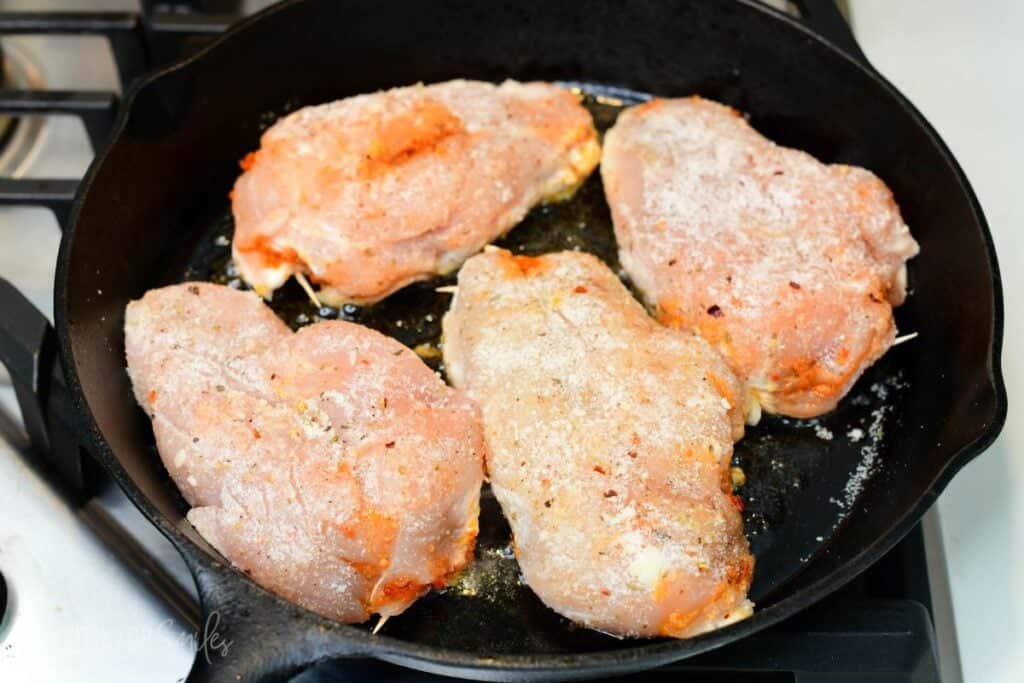 Parmesan coated stuffed chicken breasts searing in a cast iron skillet before baking.