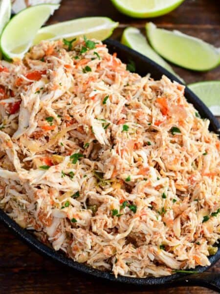 Crockpot salsa chicken with shredded chicken, peppers, and herbs in a cast iron skillet surrounded by lime wedges.