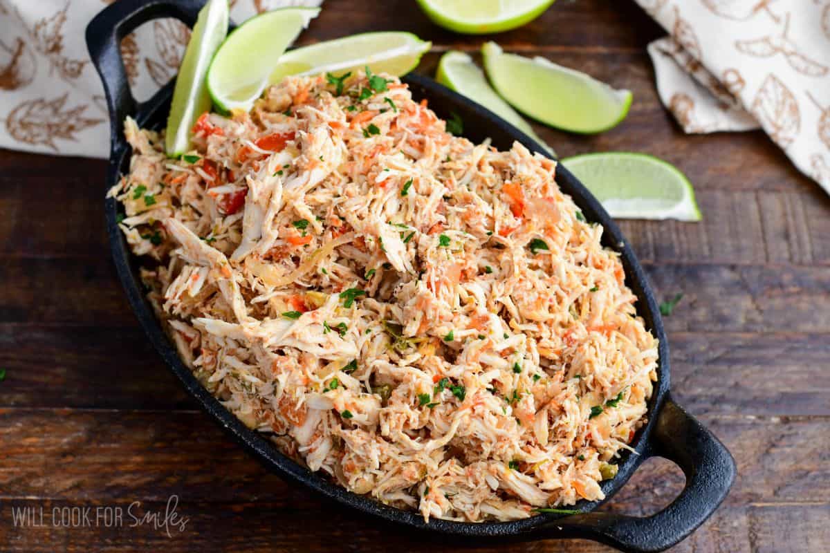 Crockpot salsa chicken with shredded chicken, peppers, and herbs in a cast iron skillet surrounded by lime wedges.