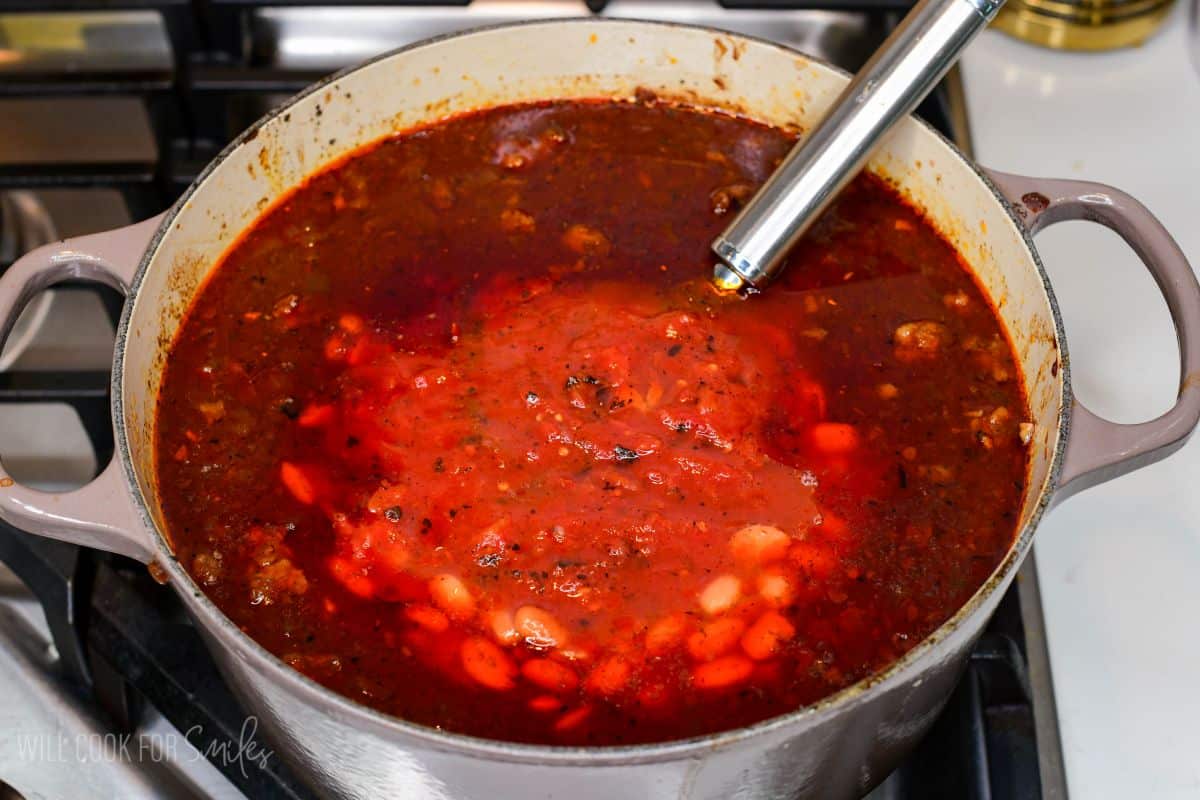 Beans and tomatoes being added to homemade short rib chili in a Dutch oven.