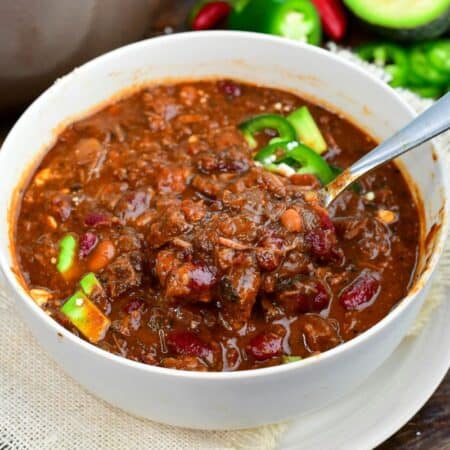 Short rib chili with tender beef and beans being scooped from a bowl topped with avocado and jalapeños.