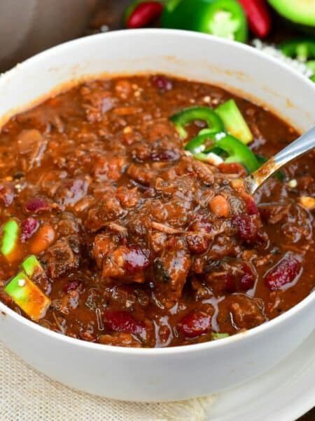 Short rib chili with tender beef and beans being scooped from a bowl topped with avocado and jalapeños.