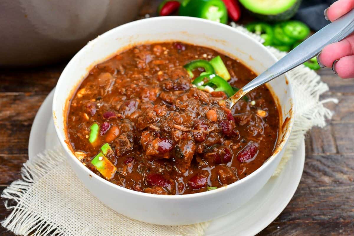 Short rib chili with tender beef and beans being scooped from a bowl topped with avocado and jalapeños.