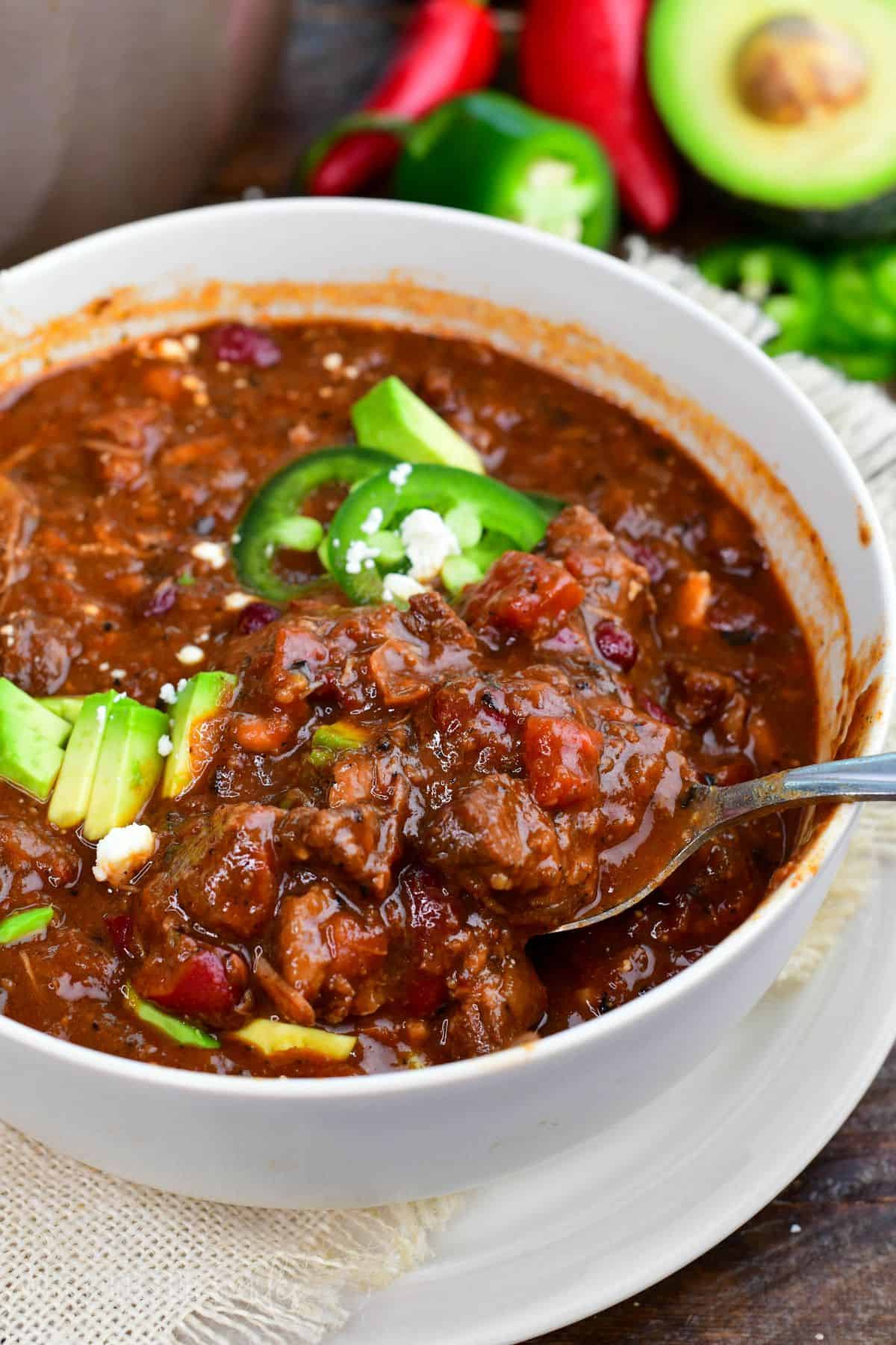 Short rib chili with tender beef, beans, avocado, and jalapeños being scooped from a bowl.